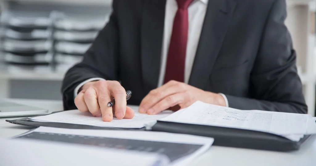 Reviewer in a suit working through printed loan data pages with a pen at an office desk, a second stack of printouts beside them